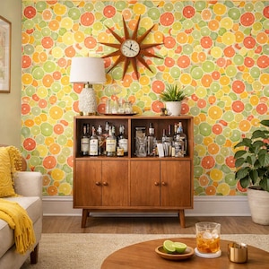 May include: A mid-century modern home bar with a wooden cabinet, a starburst clock, and a citrus fruit wallpaper. The bar is stocked with various bottles and glassware. A round wooden table holds a drink and a small bowl with lime slices.