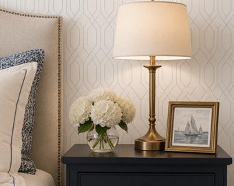 May include: A bedside table with a brass lamp, a framed picture of sailboats, and a vase of white hydrangeas. The table is dark blue, and the lamp has a beige lampshade. The background features a geometric patterned wallpaper.