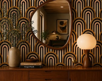 May include: An asymmetrical wooden-framed mirror hangs on a wall with black, gold, and cream arched wallpaper. Below, a wooden sideboard holds a beige vase with greenery, two stacked books titled Houses Atelier AM and Georgia O'Keeffe, a brass bird, and a lamp.