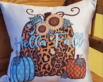 May include: A white throw pillow with a leopard print pumpkin design and the text "Hello Fall" in blue script. The pumpkin is decorated with sunflowers and blue accents.