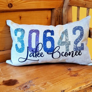 May include: A white throw pillow with a blue and purple distressed font design that reads "30642 Lake Conroe". The pillow is sitting on a wooden bench.