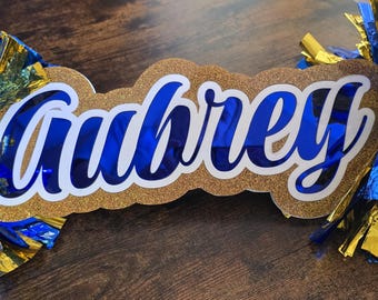 Op de afbeelding: Een gepersonaliseerd naambord met de naam Aubrey in blauwe schrijfletters met een witte omlijning, gemonteerd op een gouden glitterachtergrond, geflankeerd door twee blauw-gouden cheerleading pompons met lange, dunne, reflecterende stroken.