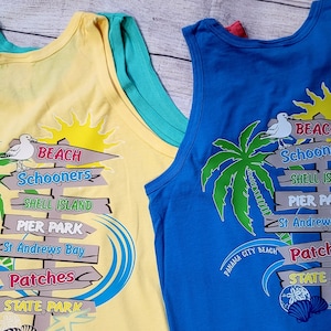 May include: Two sleeveless shirts with a beach theme. The shirts are yellow and blue with a palm tree, a seagull, and a signpost with the words "BEACH Schooners", "SHELL ISLAND", "PIER PARK", "St Andrews Bay", "Patches", and "STATE PARK".