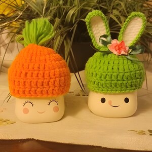 Könnte beinhalten: Zwei Keramik-Pilzfiguren mit lächelnden Gesichtern. Ein Pilz trägt einen orangefarbenen Häkelhut und der andere einen grünen Häkelhut mit Hasenohren und einer rosa Blume.