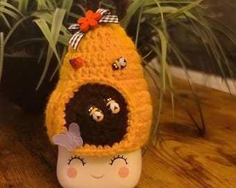 May include: A crocheted beehive with a smiling face, decorated with a butterfly, flowers, and bees. The beehive is made of yellow and brown yarn and is sitting on a wooden surface.