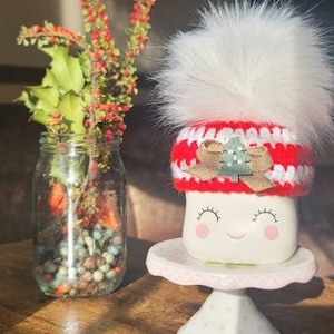 K&ouml;nnte beinhalten: Wei&szlig;e Keramiktasse mit einer rot-wei&szlig;en H&auml;kelm&uuml;tze und einem wei&szlig;en Bommel. Die Tasse hat ein l&auml;chelndes Gesicht mit rosa Wangen und Wimpern. Die Tasse steht auf einem rosa-wei&szlig;en Kuchenst&auml;nder.
