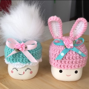 Könnte beinhalten: Zwei weiße Keramikfiguren in Marshmallow-Form mit bemalten Gesichtern, die gestrickte Mützen tragen. Eine Mütze ist rosa mit weißen Punkten und Hasenohren, die andere ist türkis mit einem weißen Bommel und einer rosa Schleife.