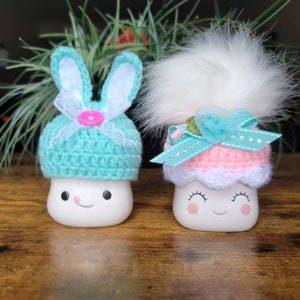 Könnte beinhalten: Zwei weiße Keramikfiguren in Marshmallow-Form mit bemalten Gesichtern. Eine Figur trägt einen hellblauen gehäkelten Hasenhut mit einer rosa Schleife. Die andere Figur trägt einen rosa gehäkelten Hut mit einem weißen gehäkelten Besatz und einer hellblauen Schleife mit einer weißen Blume.