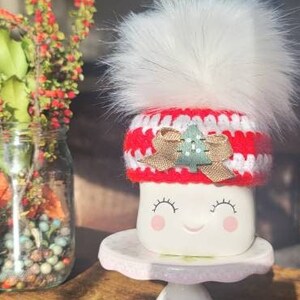 K&ouml;nnte beinhalten: Wei&szlig;e Keramiktasse mit einer rot-wei&szlig;en H&auml;kelm&uuml;tze mit einem gr&uuml;nen Baum-Applikationen und einem wei&szlig;en Bommel. Die Tasse hat ein l&auml;chelndes Gesicht mit geschlossenen Augen und rosa Wangen.