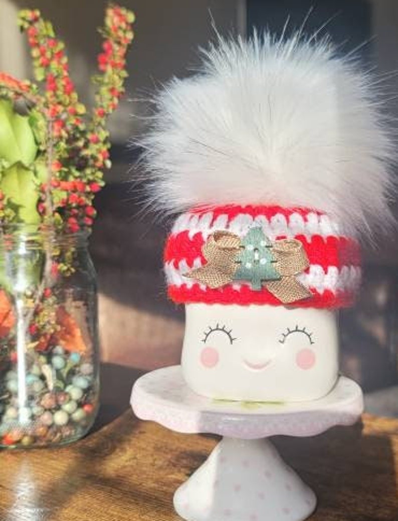 K&ouml;nnte beinhalten: Wei&szlig;e Keramiktasse mit einer rot-wei&szlig;en H&auml;kelm&uuml;tze mit einem gr&uuml;nen Baum-Applikationen und einem wei&szlig;en Bommel. Die Tasse hat ein l&auml;chelndes Gesicht mit geschlossenen Augen und rosa Wangen.