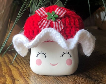 May include: A white ceramic mug with a smiling face, wearing a red crocheted hat with a white crocheted ruffle and a red and white gingham bow with a green flower.