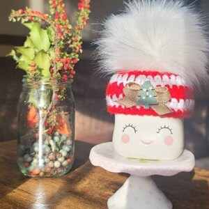 K&ouml;nnte beinhalten: Wei&szlig;e Keramiktasse mit einem l&auml;chelnden Gesicht, die einen rot-wei&szlig;en H&auml;kelhut mit einem gr&uuml;nen Weihnachtsbaum und einem wei&szlig;en Bommel tr&auml;gt. Die Tasse steht auf einem wei&szlig;en Kuchenst&auml;nder mit rosa Punkten.