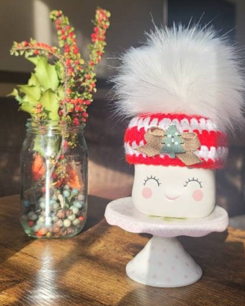 K&ouml;nnte beinhalten: Wei&szlig;e Keramiktasse mit einem l&auml;chelnden Gesicht, die einen rot-wei&szlig;en H&auml;kelhut mit einem gr&uuml;nen Weihnachtsbaum und einem wei&szlig;en Bommel tr&auml;gt. Die Tasse steht auf einem wei&szlig;en Kuchenst&auml;nder mit rosa Punkten.