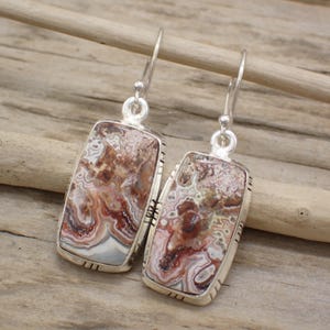 May include: A pair of rectangular dangle earrings featuring polished Crazy Lace Agate stones set in sterling silver frames. Each earring has a silver hook wire and displays intricate, swirling patterns of brown, cream, and reddish-orange colors.