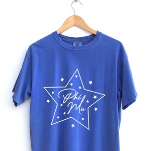 Puede incluir: Una camiseta azul con un diseño de estrella blanca y el texto "Phi Mu" dentro de la estrella.