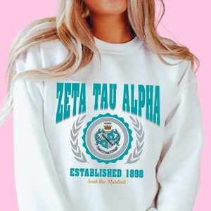 Może przedstawiać: Biała bluza z nadrukiem graficznym w kolorze turkusowym i szarym z napisem "Zeta Tau Alpha Established 1898 Seek the Noblest".