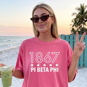 Könnte beinhalten: Ein korallenrotes Kurzarm-T-Shirt mit weißem Siebdrucktext 1867 PI BETA PHI und fünf kleinen Sternen, die zwischen den Zahlen und dem Text zentriert sind. Das T-Shirt wird mit hellblauen Jeansshorts kombiniert.