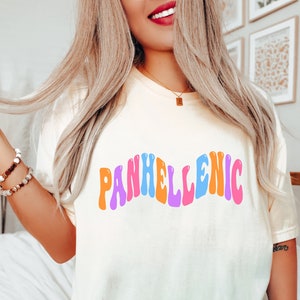 Puede incluir: Camiseta color crema con la palabra "PANHELLENIC" en una fuente colorida y ondulada. El texto es en tonos naranja, rosa, azul y morado. La camiseta es de estilo casual.