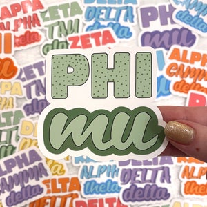 Puede incluir: Un primer plano de una pegatina verde y blanca con el texto "PHI mu" en una fuente manuscrita en negrita. La pegatina está rodeada de otras pegatinas con varias letras y nombres griegos.