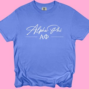 Puede incluir: Una camiseta azul bígaro con las palabras "Alpha Phi" en escritura blanca en la parte delantera. Debajo de la escritura están las letras griegas "AΦ" en blanco. La camiseta está hecha de un material suave y cómodo.