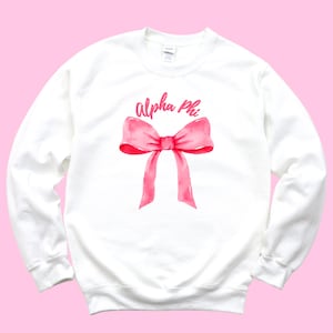 Puede incluir: Sudadera blanca con un diseño de acuarela de un lazo rosa y las palabras "alpha phi" en escritura rosa. La sudadera de manga larga tiene cuello redondo. El diseño está centrado en la parte delantera de la prenda.
