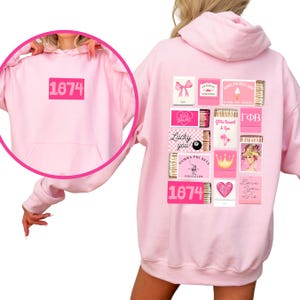 May include: Pink hoodie with a graphic print of matchboxes with various designs, including a crown, a heart, a palm tree, and the text "Lucky you!" and "Gamma Phi Beta Polo Club". The hoodie also has the number "1874" printed on the front and back.