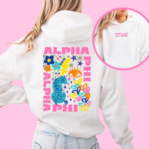 May include: White hoodie featuring a vibrant graphic on the back. The design incorporates the words "ALPHA PHI" in pink, a blue and turquoise tiger, flowers, stars, and a lightning bolt. The front of the hoodie displays the text "alpha phi est. 1872" in pink.