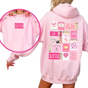 May include: A light pink hoodie with a front pocket and a hood. The front features the number "1872" in a pink rectangle. The back displays various pink graphic designs, including matchbox-style illustrations with text and symbols.