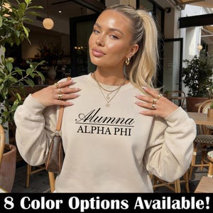 Op de afbeelding: Een beige sweatshirt met de tekst "Alumna ALPHA PHII" in zwart script. De afbeelding toont een vrouw die het sweatshirt draagt, geaccessoireerd met gouden sieraden. De onderkant van de afbeelding heeft de tekst "8 Color Options Available!"