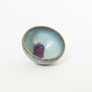 May include: A small, handmade ceramic bowl with a rounded shape. The interior is a blend of blue and teal hues, with a deep purple accent. The rim has a brown, textured finish. The bowl is set against a white background.