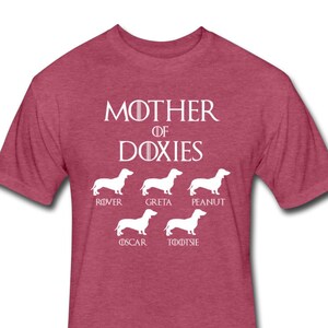May include: A heather red t-shirt with the text "Mother of Doxies" in white. Below the text are five white silhouettes of Dachshunds with the names "Rover", "Greta", "Peanut", "Oscar", and "Tootsie" underneath each dog.