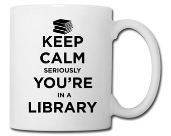 Puede incluir: Taza de café de cerámica blanca con texto negro que dice "Keep Calm Seriously You're In A Library". Una pila de tres libros está representada encima del texto.