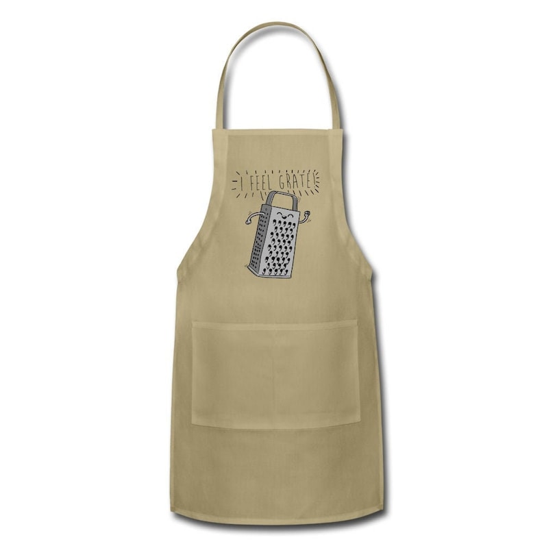Pu&ograve; includere: Un grembiule da cucina beige con una tasca presenta un'illustrazione di un grattugia con un viso sorridente e il testo "I feel grate".