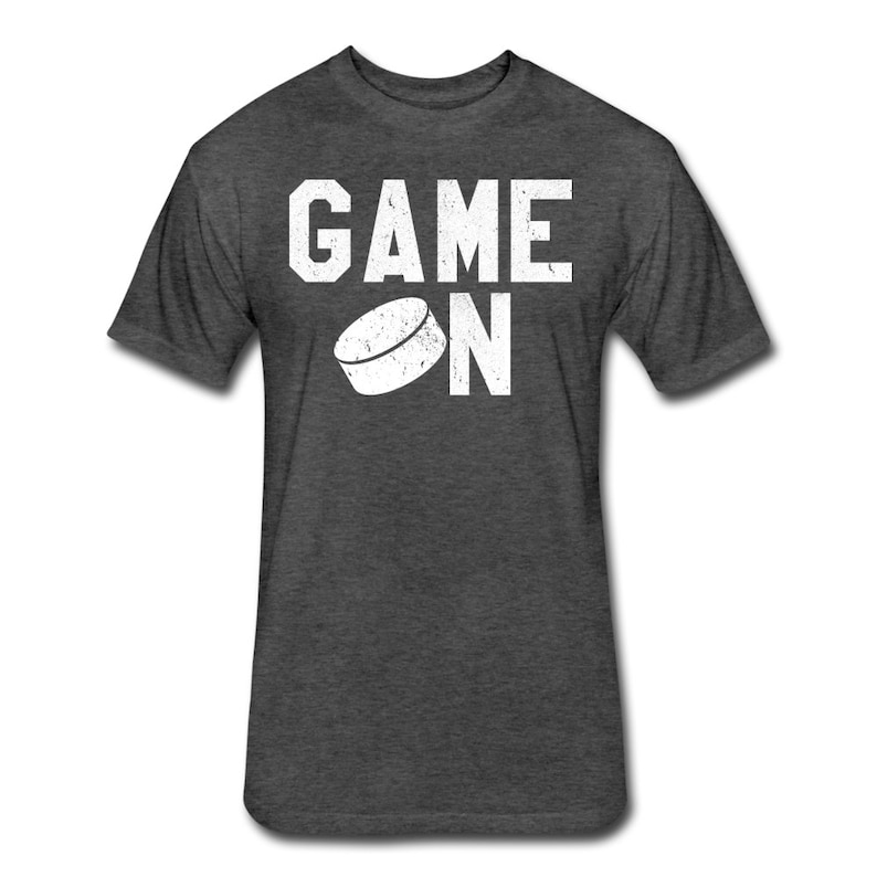 May include: A dark heather grey t-shirt with a white graphic of a hockey puck and the text "GAME ON".
