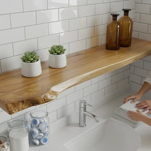 Peut inclure: Une étagère en bois à bord brut avec deux petites plantes succulentes en pot et deux bouteilles en verre ambré. Une femme et un enfant plient des serviettes blanches dans une salle de bain carrelée de blanc.