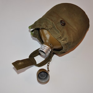May include: A green canvas canteen with a metal lid and a chain attached. The canteen is closed and has a brown button on the top.