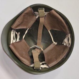 May include: An olive green military helmet, viewed from the inside. It features a brown leather liner with multiple pads and a central strap. White cords secure the liner to the helmet's interior. The helmet is designed for head protection.