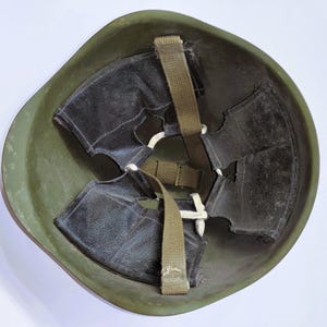 Puede incluir: Un casco militar verde oliva, visto desde el interior. El casco presenta un interior oscuro con acolchado negro y un sistema de correas color caqui. El acolchado interior está sujeto con hilo blanco.