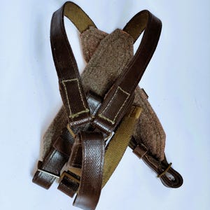 May include: Brown leather and canvas suspenders with a felted wool shoulder pad. The suspenders have adjustable straps and metal hardware. The overall color scheme is brown and olive green.
