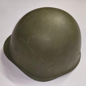 Puede incluir: Un casco militar verde oliva con una corona redondeada y un borde. El casco está hecho de un material duradero y tiene una superficie ligeramente texturizada. El casco está diseñado para proteger la cabeza del usuario. Es un equipo militar.