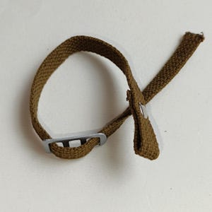以下が含まれることがあります： 銀色の金属製のバックルが付いた茶色のキャンバスストラップ。ストラップの一方の端にはループがあり、もう一方の端には小さな金属製のボタンが付いています。