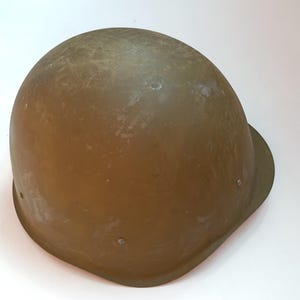 Puede incluir: Un casco militar de color marrón claro con una forma ligeramente redondeada y una superficie lisa. El casco tiene varios pequeños remaches metálicos alrededor de los bordes.