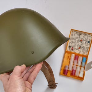 May include: An olive green military helmet with a leather chin strap sits next to an open orange first aid kit. The kit contains several small vials of various colors. The helmet has a smooth, matte finish.