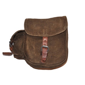 Puede incluir: Una bolsa de lona marrón con una correa de cuero y una hebilla. La bolsa tiene una solapa que se cierra con una hebilla.