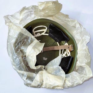 Puede incluir: Un casco militar verde oliva con una correa de barbilla de cuero y acolchado interior negro. El casco está parcialmente envuelto en papel translúcido y arrugado. Se ve una cuerda blanca dentro del casco.