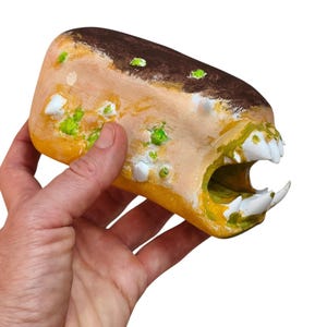 May include: A hand-painted, oblong creature figurine featuring a tan and brown body with textured green spots, white protruding spikes, and an open mouth displaying sharp, pointed white teeth. The object has a smooth, rounded top surface.