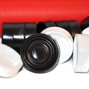 Puede incluir: Un juego de damas negras y blancas, algunas apiladas unas sobre otras. Las damas son de plástico y tienen un acabado liso y brillante.