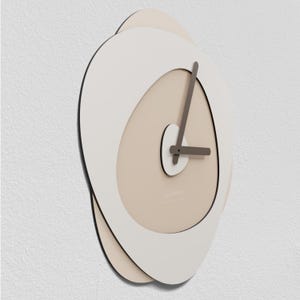 Op de afbeelding: Een moderne wandklok met een uniek, gelaagd ontwerp. De klok heeft een wit, ovaalvormig frame met een beige interieur en donkergrijze wijzers. De klok is aan een witte muur bevestigd.