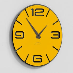 Può includere: Un orologio da parete moderno con un quadrante giallo brillante e numeri neri alle 3, 6, 9 e 12. Le lancette dell'orologio sono grigio scuro e l'orologio ha un bordo nero. L'orologio è montato su una parete bianca.