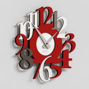 Peut inclure: Une horloge murale moderne avec un schéma de couleurs rouge, blanc et gris. L'horloge présente de grands chiffres stylisés en rouge et blanc, avec un centre circulaire et des aiguilles argentées. L'horloge est marquée "Made in Italy".